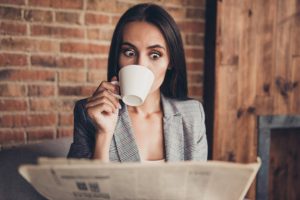 WOW! Close up portrait of astonishment brunette woman with crazy emotion and stare open eyed read the news about high oil prices drink espresso beverage from a white small mug in loft workstation