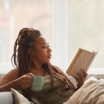 African young woman lying on sofa drinking coffee and reading an interesting book at home
